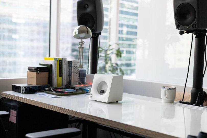 Small square-shaped white audio device with small circular window containing a globule of nanoscale ferromagnetic particles designed to move in relation to music set own top of a glossy white table top desk, surrounded by a pair of monitor speakers, desk lamp, books and other indeterminate objects. Backdrop of large windows in background.
