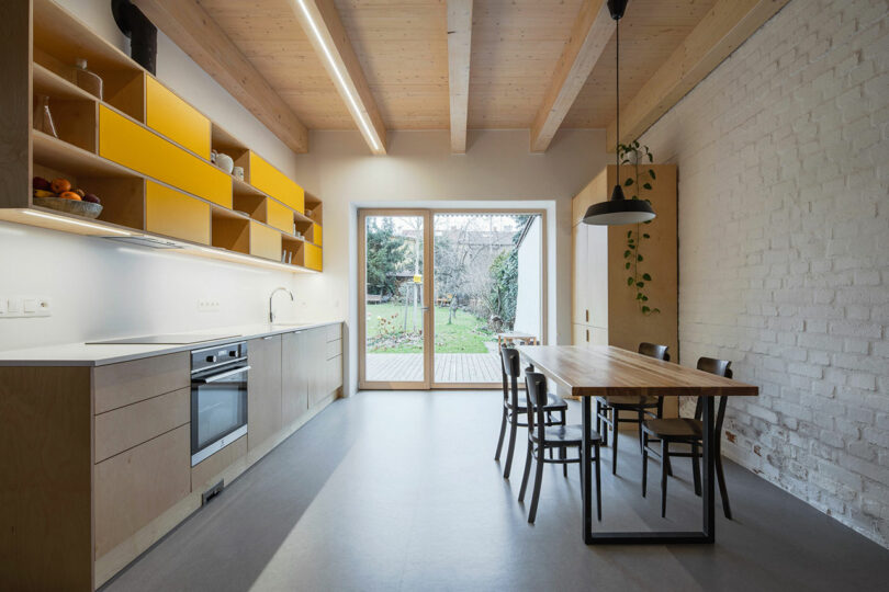 long interior view of modern minimalist kitchen with dining table to right and wood and yellow cabinets to left