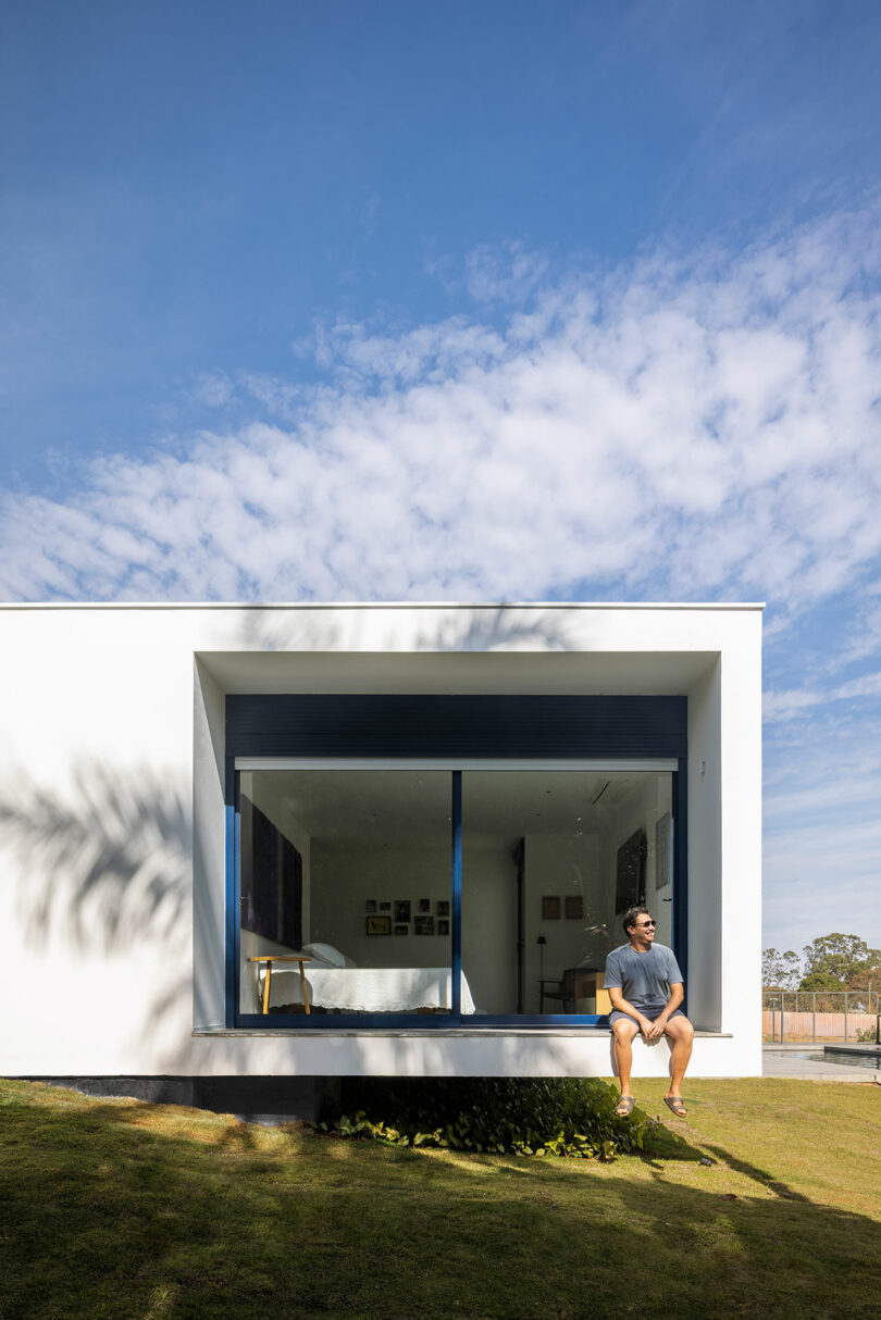 partial exterior view of modern white house with bedroom that opens to an outdoor terrance with man sitting on ledge