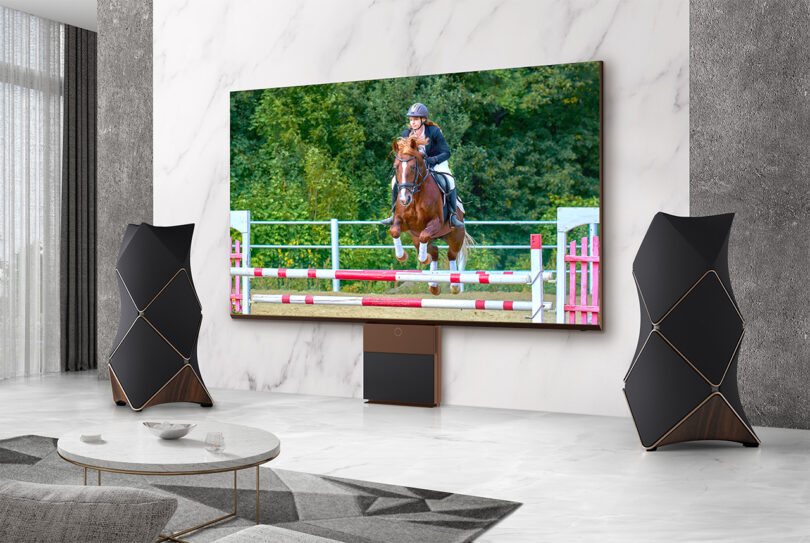 LG MAGNIT 136-inch Micro LED television in a modern living room setting flanked on both sides by the tessellated 4-foot tall Beolab 90 floor speakers. On the screen is a young woman on horseback participating in equestrian.