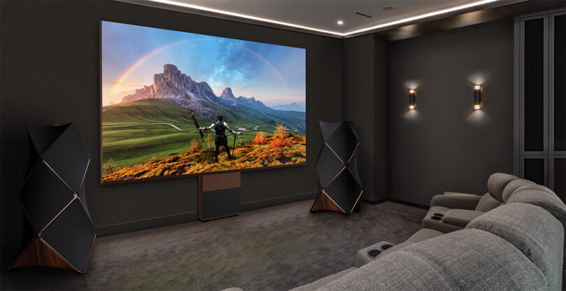 LG MAGNIT 136-inch Micro LED television in darkened home theater setting flanked on both sides by the tessellated 4-foot tall Beolab 90 floor speakers. On the screen is a fantasy fiction animation or video game, with a wondrous landscape with arching rainbow over dramatic mountaintops.