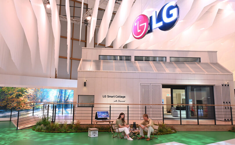 Exterior of LG Smart Cottage showcase home at IFA 2023 with man and woman seated in chairs on faux grass. Large LG logo hangs overhead.